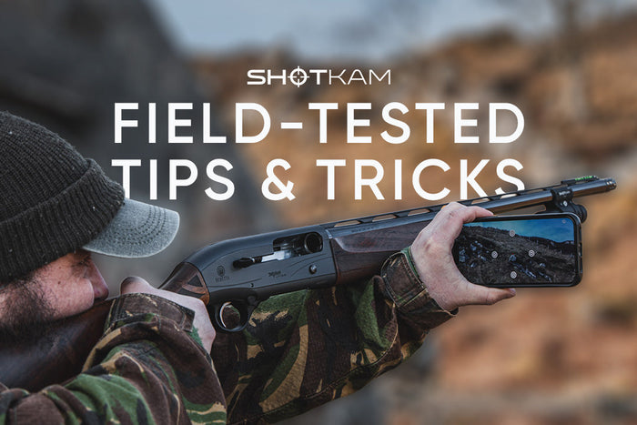 Shooter using a ShotKam barrel-mounted camera on a shotgun, checking recorded target footage on a mobile phone outdoors.
